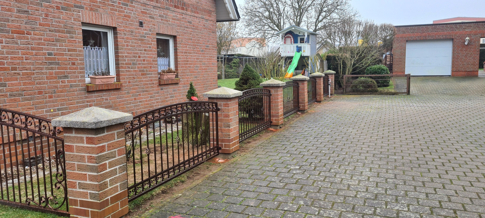 Kute ogrodzenie z ceglanymi słupkami i betonowymi nakrywami, przylegające do ceglanego domu z oknem i skrzynką z kwiatami, w tle domek dla dzieci ze zjeżdżalnią oraz garaż.