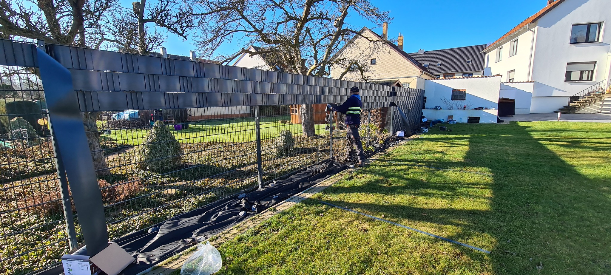 Montaż szarego ogrodzenia panelowego na metalowej konstrukcji; widoczny pracownik w odblaskowej kamizelce na tle zielonego trawnika i zabudowań.