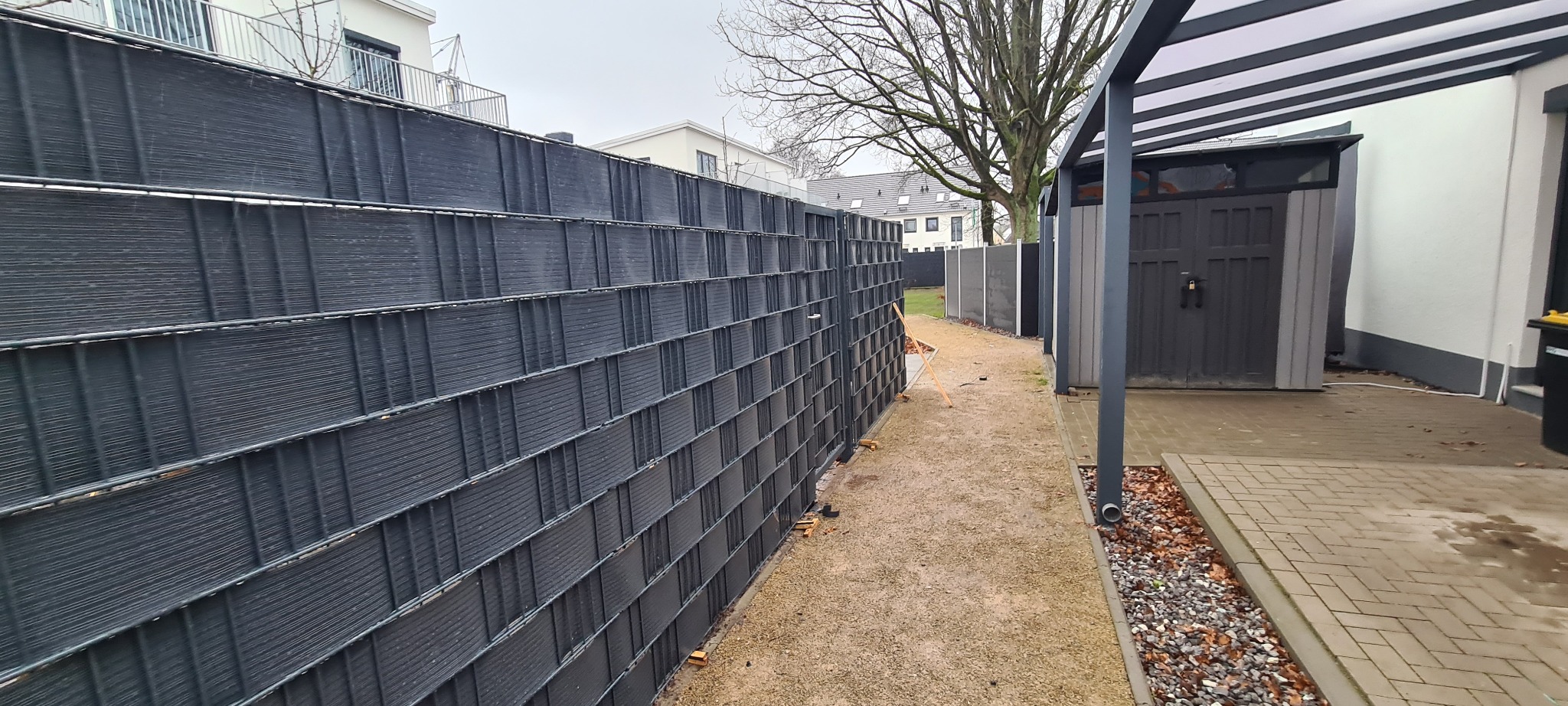 Długi, ciemny, panelowy płot w Różańsku, z widoczną konstrukcją kratową i wypełnieniem, oddzielający posesję od ścieżki z drobnym żwirem, z budynkami w tle.