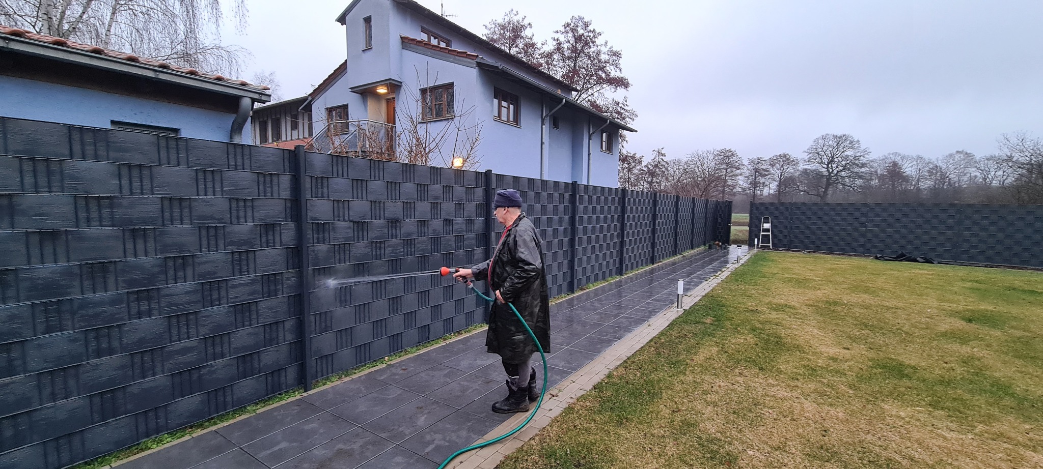 Mężczyzna w płaszczu czyści wodą pod ciśnieniem szary panelowy płot na tle domu z ogrodem w pochmurny dzień.