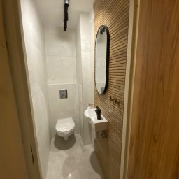 Usługi Ogólnobudowlane Zoch-Bud - Wąska, nowoczesna toaleta z drewnianą ścianą, owalnym lustrem, białą umywalką i podwieszaną miską WC. Reflektory sufitowe oświetlają wnętrze.