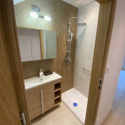 Usługi Ogólnobudowlane Zoch-Bud - Nowoczesna łazienka z kabiną prysznicową typu walk-in, umywalką z szafką w odcieniach drewna i lustrem. Ściana zdobiona geometrycznym wzorem z drewnianych paneli.