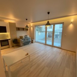 Usługi Ogólnobudowlane Zoch-Bud - Jasne wnętrze po remoncie z panelami podłogowymi i nową zabudową kuchenną. Widoczne lampy wiszące i duże okno balkonowe. Część mebli jeszcze zapakowana.