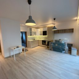 Usługi Ogólnobudowlane Zoch-Bud - Jasne panele podłogowe w nowym mieszkaniu z kuchnią w zabudowie, oświetleniem punktowym i meblami w folii. Widok na salon i kuchnię po remoncie.