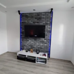 Ściana z szarej cegły z niebieskim podświetleniem LED za telewizorem. Biała szafka RTV z czarnymi szufladami. Jasna podłoga. Nowoczesny wystrój wnętrza.