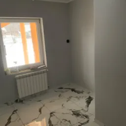 Wnętrze pokoju po remoncie: świeżo pomalowane szare ściany, marmurowe płytki podłogowe z ciemnymi żyłami, biały grzejnik pod oknem z widokiem na zimowy krajobraz.