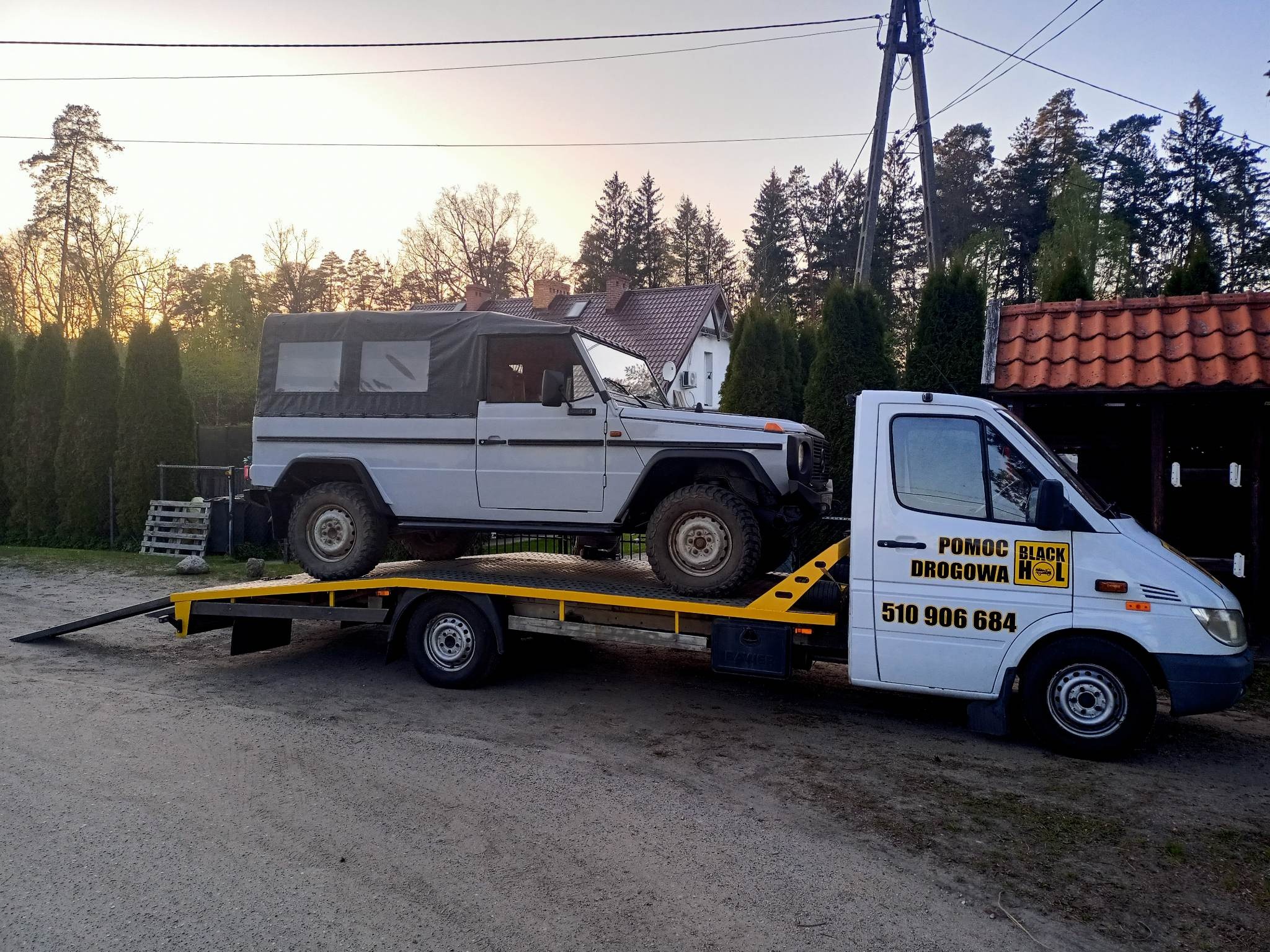 Laweta z Olsztyna przewozi terenowy samochód z brezentowym dachem na tle domów i drzew o zmierzchu.