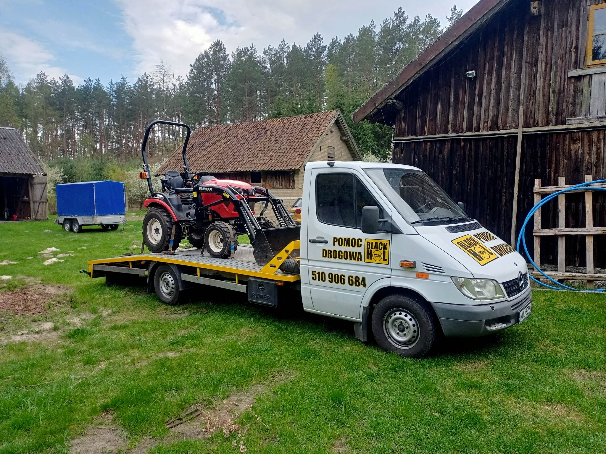 Laweta z czerwonym traktorem Yanmar na platformie, zaparkowana na trawniku przed starym drewnianym budynkiem gospodarczym, widoczna przyczepa z plandeką.
