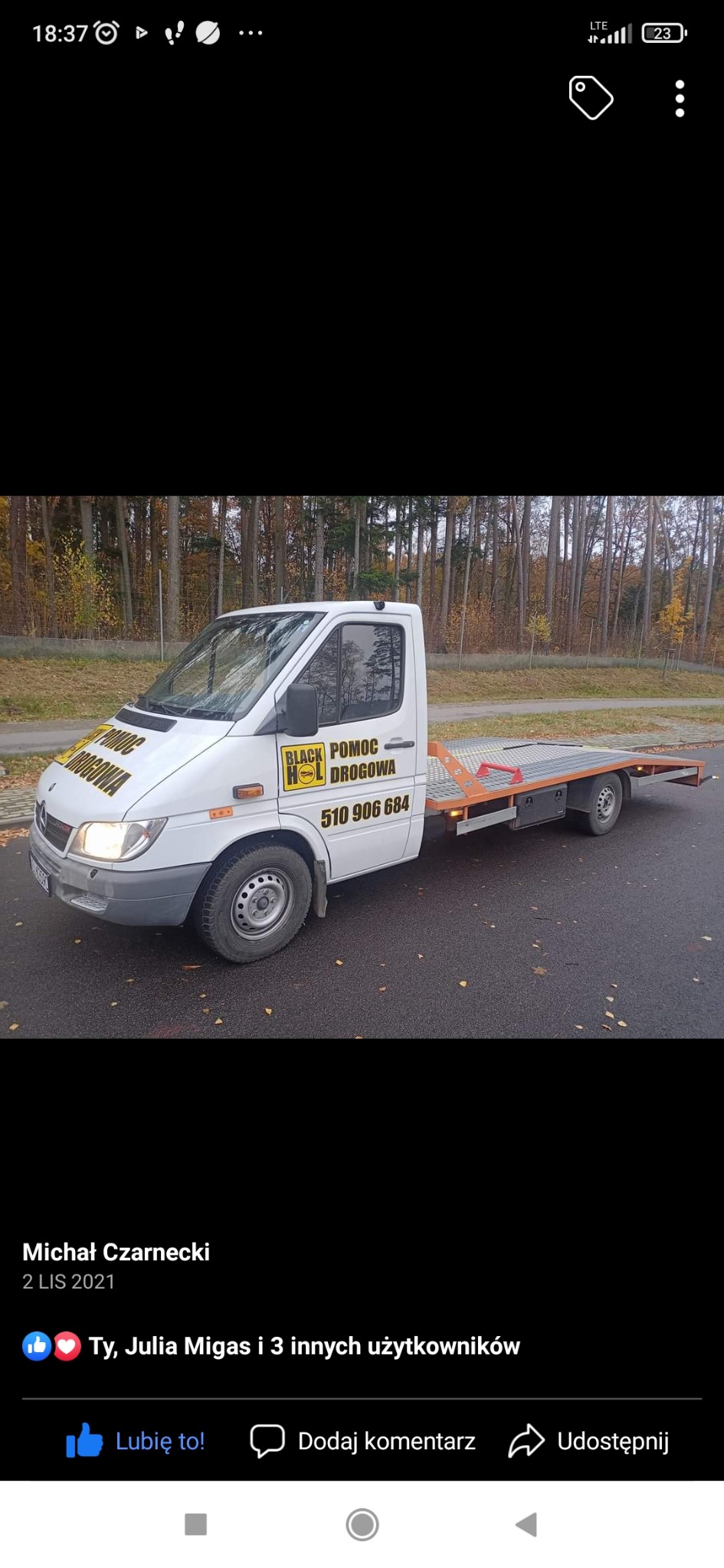 Biała laweta Mercedes z pomarańczową platformą transportową zaparkowana na asfalcie, z logo 'Black Hol Pomoc Drogowa' i numerem telefonu na boku kabiny, jesienne drzewa w tle.