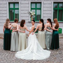 Panna młoda w białej sukni ślubnej z długim trenem, trzymająca bukiet białych kwiatów, otoczona przez druhny w zielonych i srebrnych sukniach na tle budynku z zielonymi oknami i brukowanej ulicy.