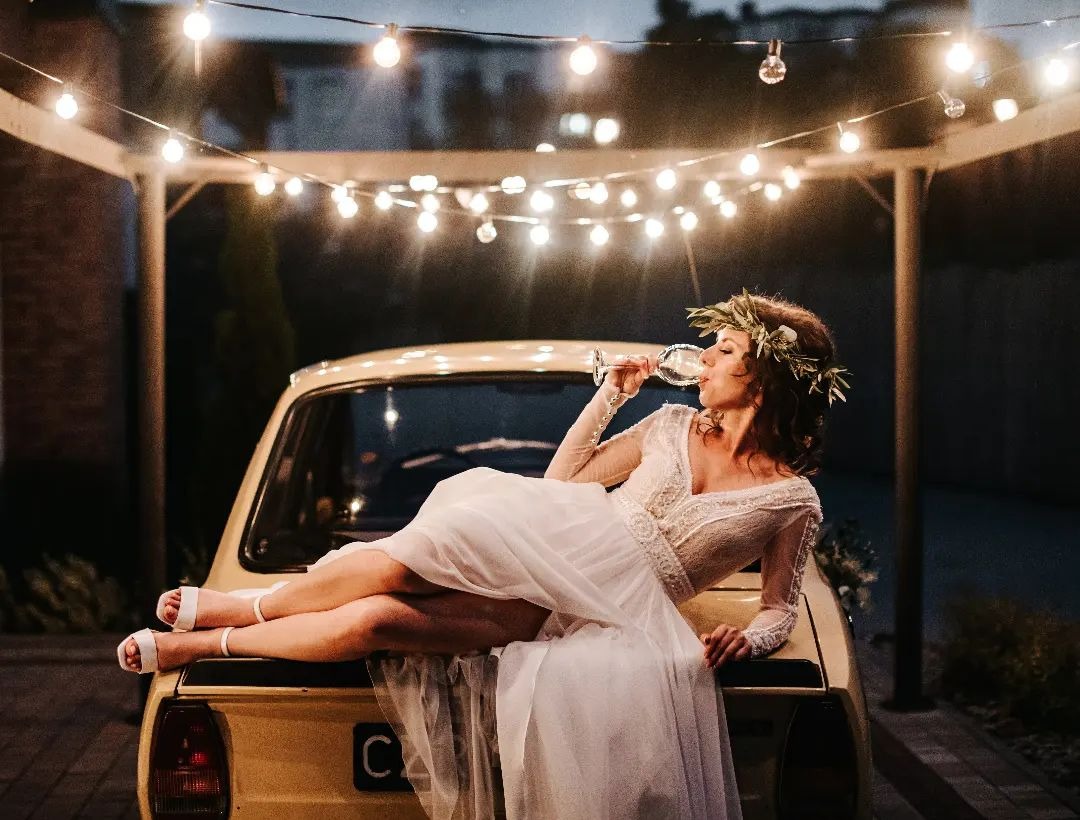 Panna młoda w koronie z liści pije wino leżąc na żółtym samochodzie, oświetlonym girlandą żarówek.