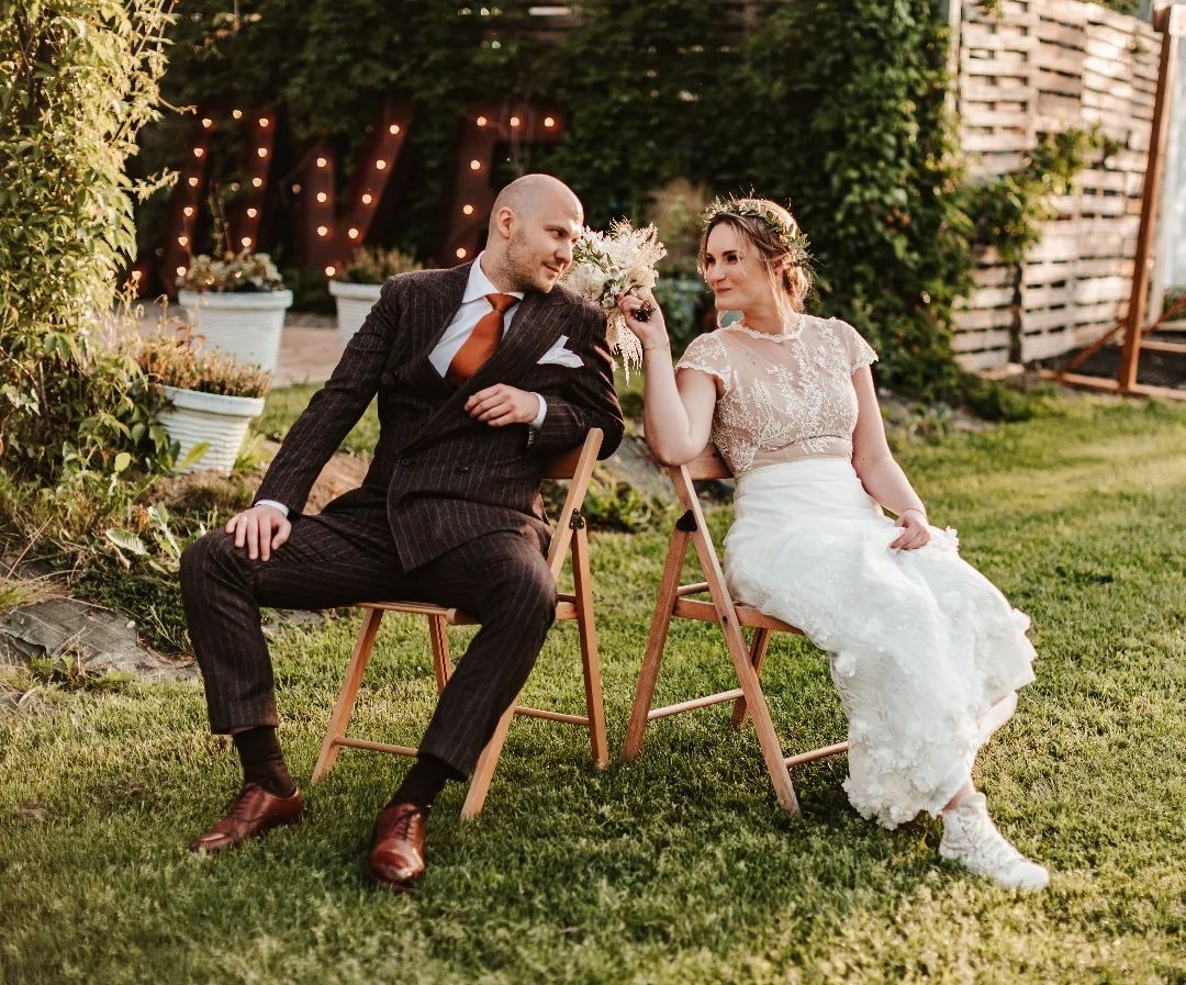 Para młoda siedząca na drewnianych krzesłach na trawie, on w brązowym, prążkowanym garniturze z pomarańczowym krawatem, ona w koronkowej sukni i wianku, w tle dekoracje świetlne i drewniana...