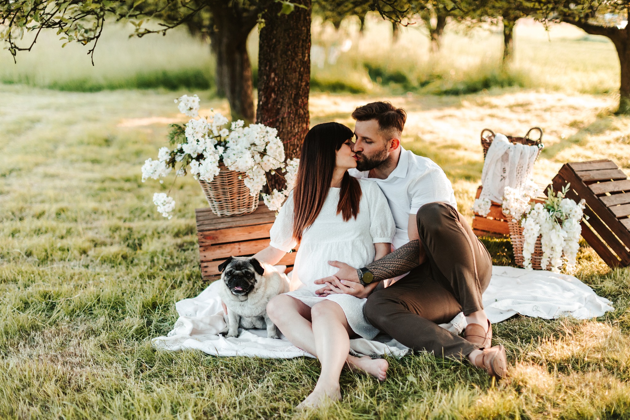 Kobieta w białej sukience w ciąży i mężczyzna w białej koszuli całują się na trawie, obok nich siedzi mops, a w tle białe kwiaty w koszyku i drewniana skrzynka.