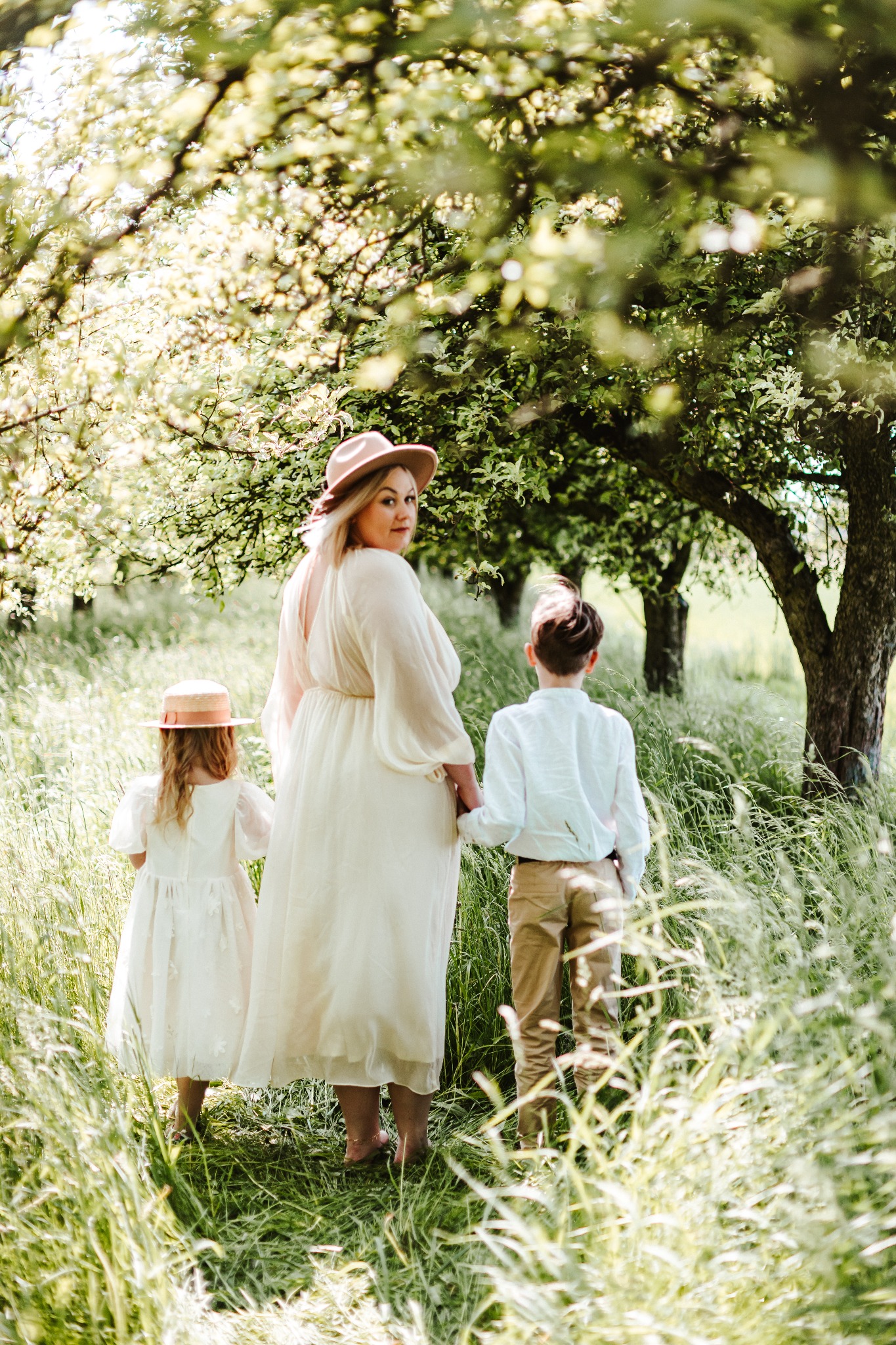 Kobieta w jasnej sukience i kapeluszu, trzymająca za ręce małą dziewczynkę w białej sukience i chłopca w koszuli i spodniach, idąca przez łąkę w słoneczny dzień, spoglądająca przez ramię.
