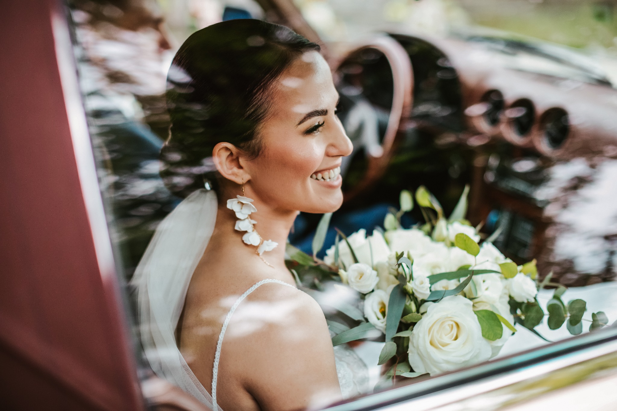 Uśmiechnięta panna młoda w samochodzie retro, widoczna przez szybę, z bukietem białych róż i eukaliptusa, ozdobne kolczyki w kształcie kwiatów.