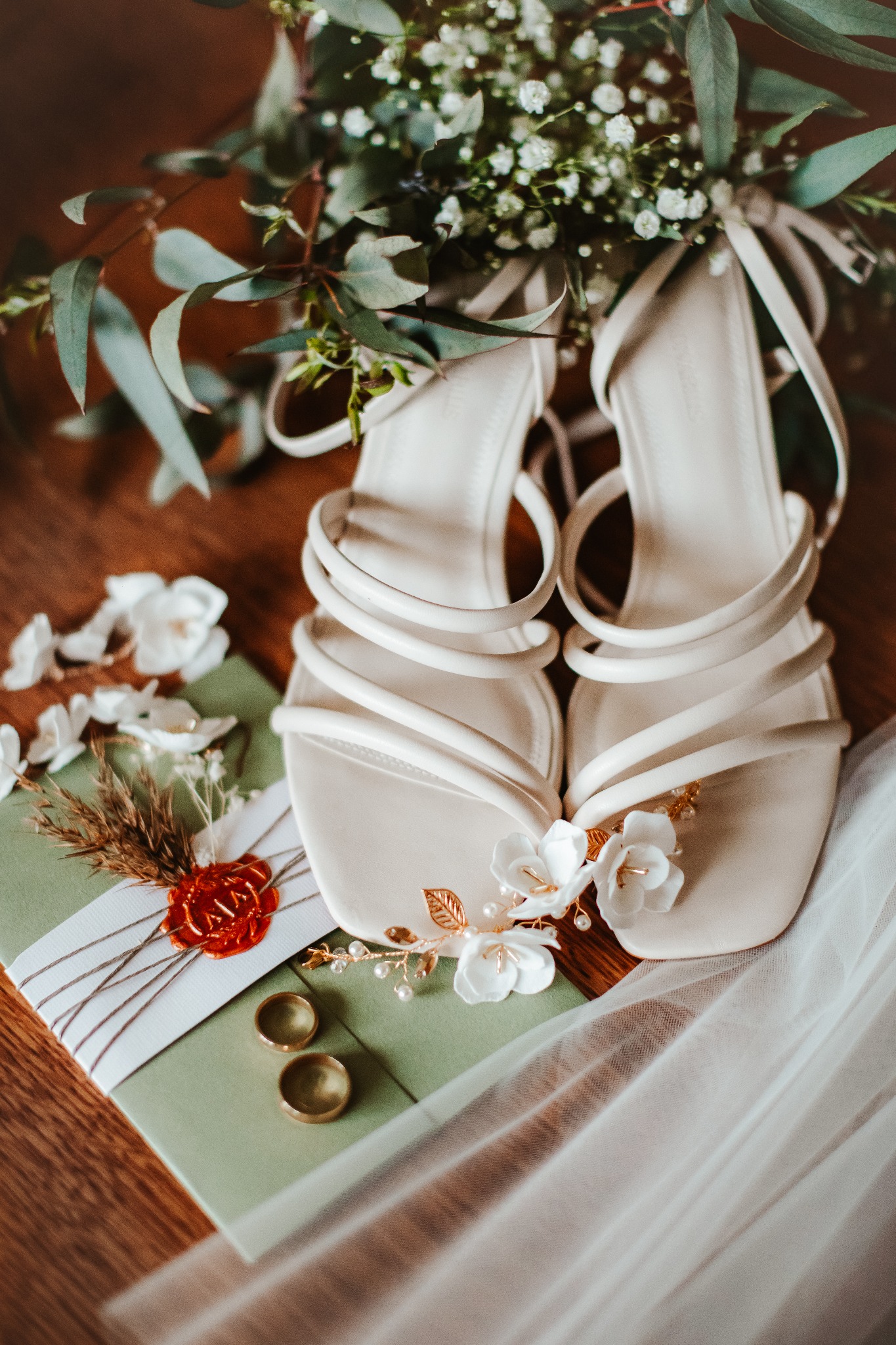 Eleganckie białe sandałki z paskami, zdobione kwiatami i złotymi listkami, leżą na zielonej kopercie z pieczęcią lakową, obok obrączki i bukiet z eukaliptusa i gipsówki, całość na drewnianym tle.