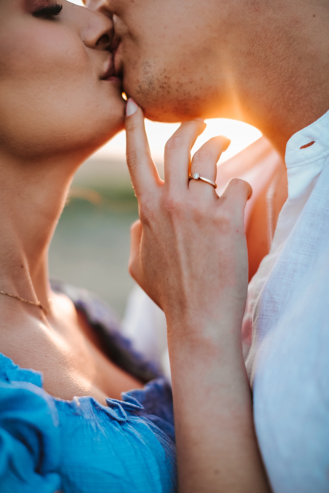 Zbliżenie na parę całującą się w kontraście ze słońcem, dłoń kobiety z pierścionkiem zaręczynowym delikatnie dotyka twarzy mężczyzny.