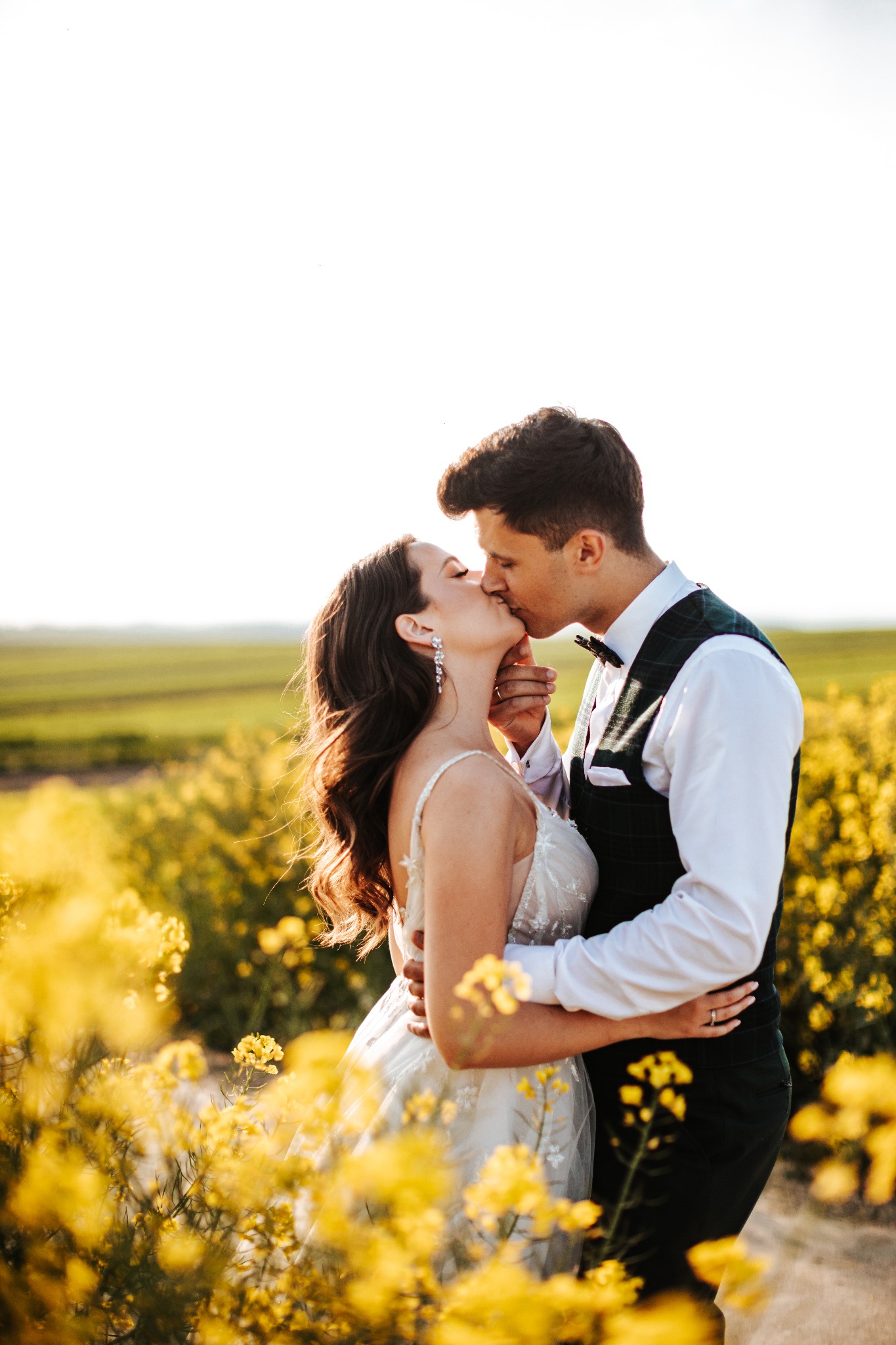 Pocałunek pary młodej w polu kwitnącego rzepaku, suknia ślubna z koronkowymi aplikacjami, zielona kamizelka pana młodego, subtelne kolczyki panny młodej.