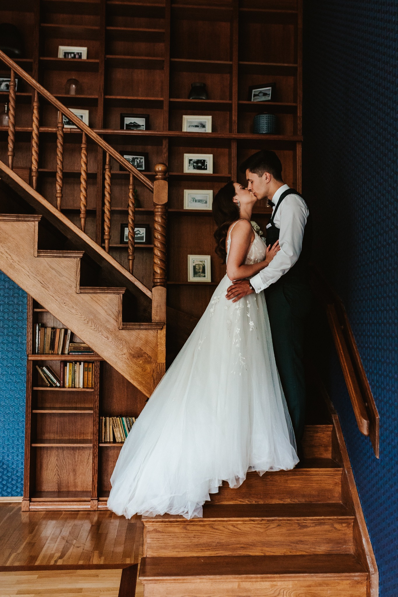 Pocałunek nowożeńców na drewnianych schodach przed regałem z książkami i dekoracjami.