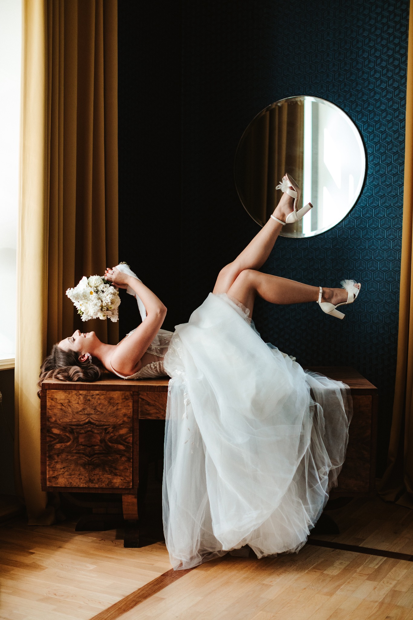 Panna młoda w białej sukni leży na drewnianej komodzie, trzyma bukiet białych kwiatów, nogi uniesione w białych butach na obcasie, w tle okrągłe lustro i wzorzysta ściana.