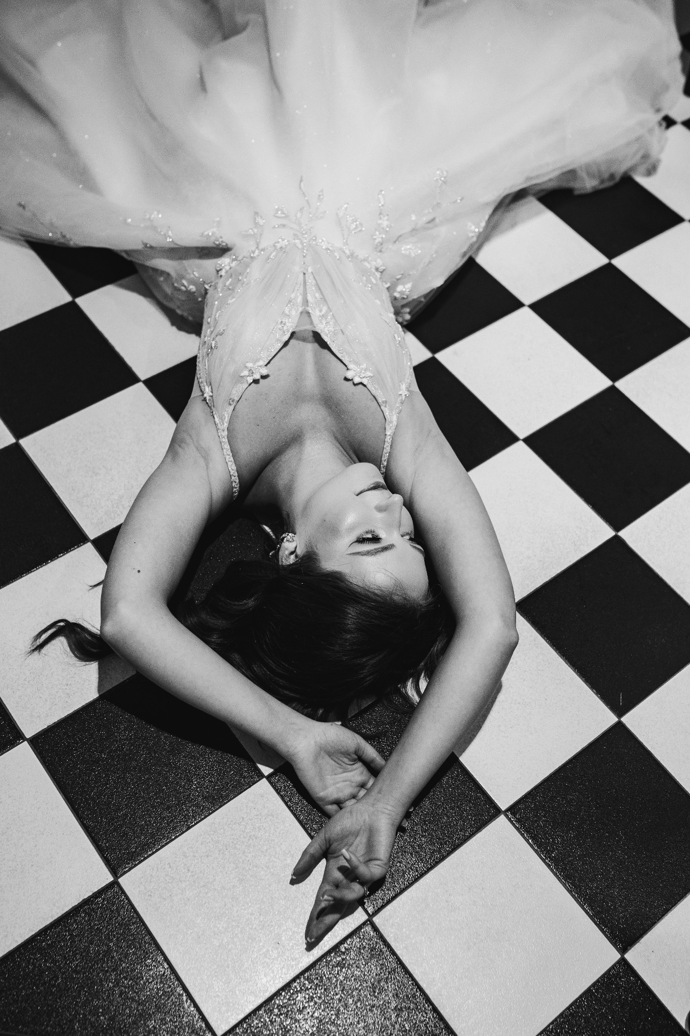 Kobieta w sukni ślubnej leżąca na czarno-białej szachownicy, ujęcie z góry, czarno-białe.