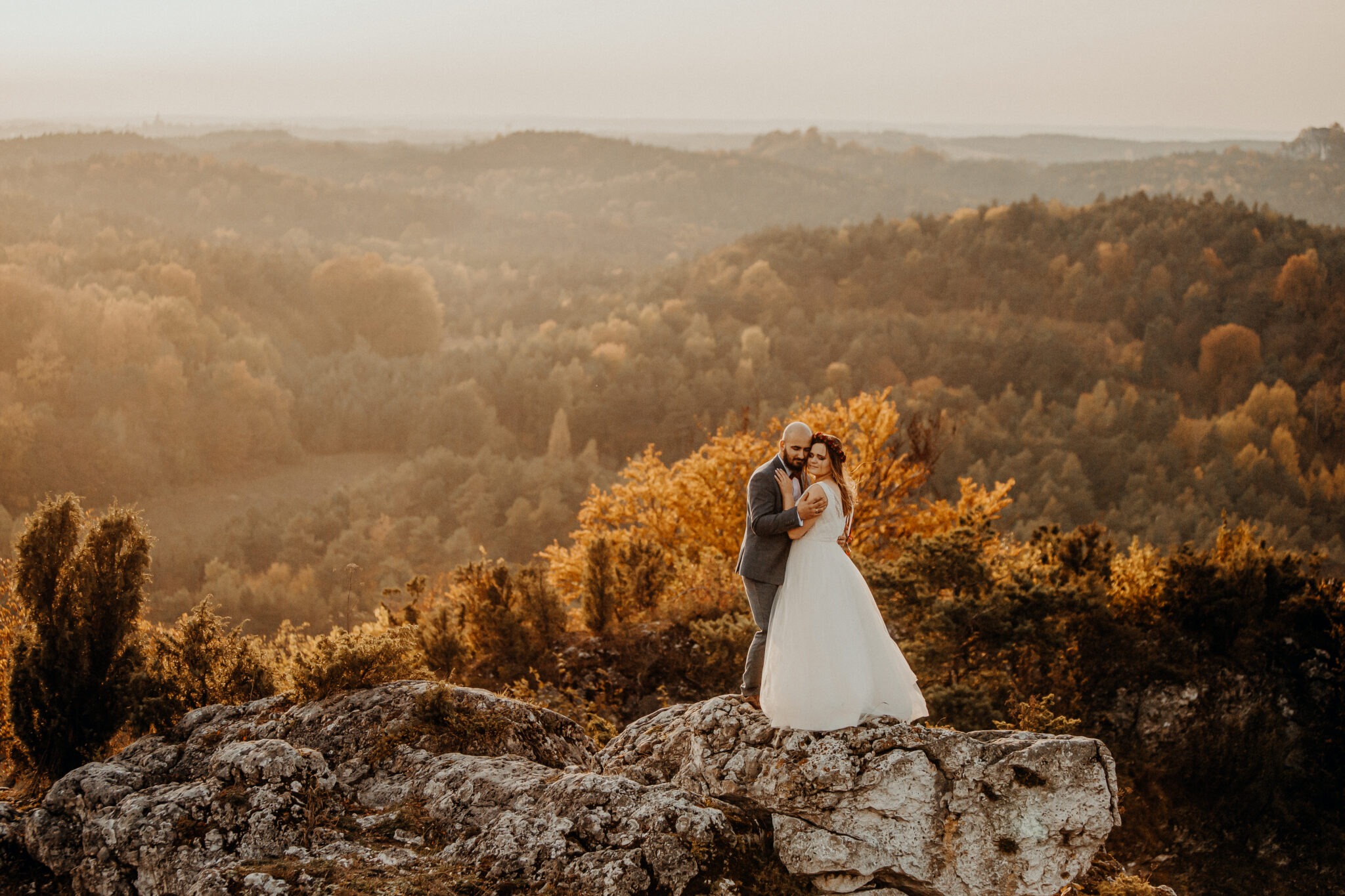 Para młoda w objęciach na skalistym wzgórzu z widokiem na zalesione doliny w ciepłym, zachodzącym słońcu.