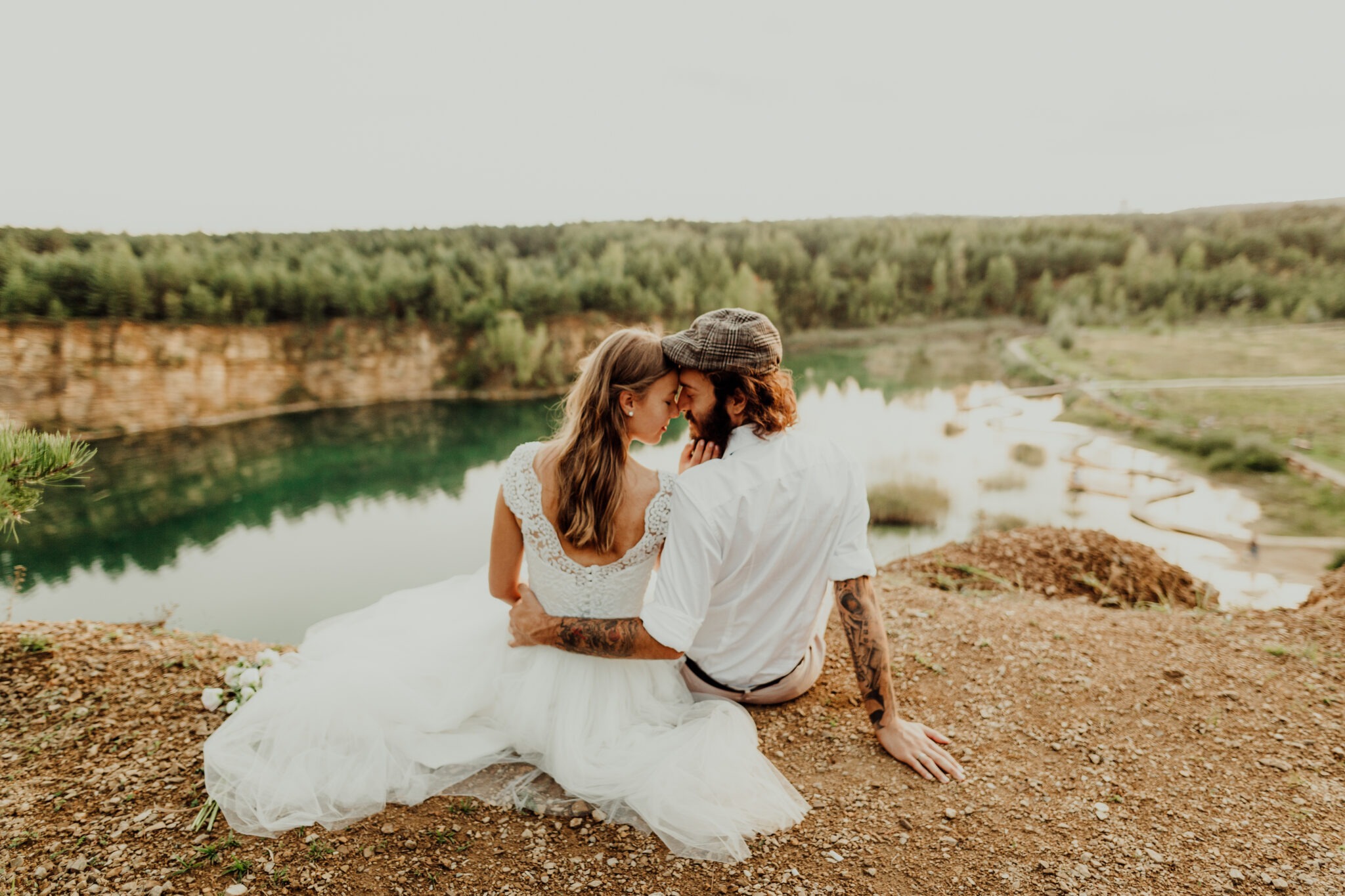 Para młoda siedzi na wzgórzu z widokiem na jezioro w dawnym kamieniołomie, panna młoda w koronkowej sukni, pan młody w kaszkiecie i z tatuażami na ramionach.