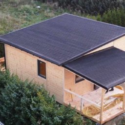 Piotr - Surowy stan domu z jasnego drewna z ciemnym dachem bitumicznym, widok z góry na budowę z zewnątrz, w tle trawa i drzewa.