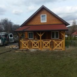 Piotr - Drewniany domek z czerwonym dachem i ażurową balustradą werandy, częściowo zasłonięty przez zaparkowany biały bus, widoczny w pochmurny dzień na tle drzew i wzgórz.