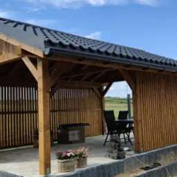 Drewniana altana z czarnym dachem, pionowe deski boczne, stół i krzesła w środku, kobieta podlewa kwiaty obok, widok na pola w tle.