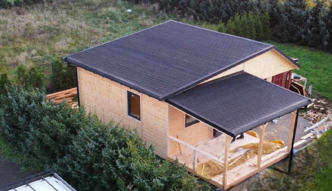 Surowy stan domu z jasnego drewna z ciemnym dachem bitumicznym, widok z góry na budowę z zewnątrz, w tle trawa i drzewa.