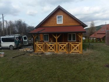 Drewniany domek z czerwonym dachem i ażurową balustradą werandy, częściowo zasłonięty przez zaparkowany biały bus, widoczny w pochmurny dzień na tle drzew i wzgórz.