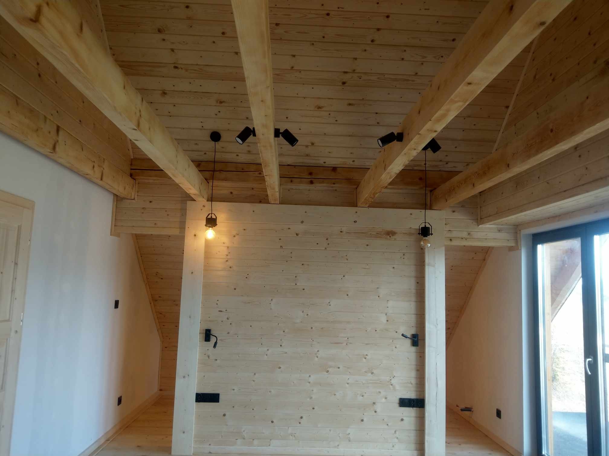 Wnętrze domu z drewnianym stropem belkowym, widoczne czarne reflektory punktowe i wiszące lampy z żarówkami Edisona, ściany wykończone boazerią.