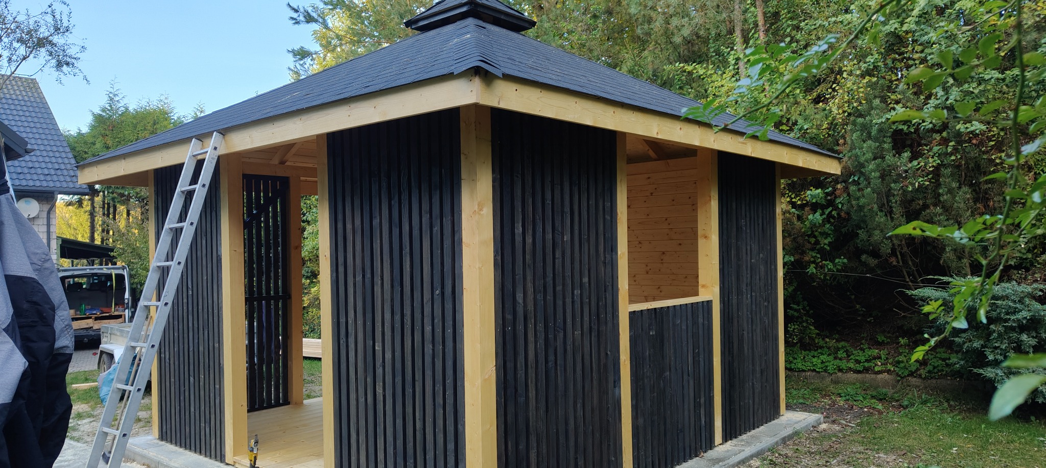 Wznoszenie drewnianej altany z czarnymi panelami pionowymi, widoczna drabina oparta o konstrukcję, elementy dachu z czarnej dachówki bitumicznej.