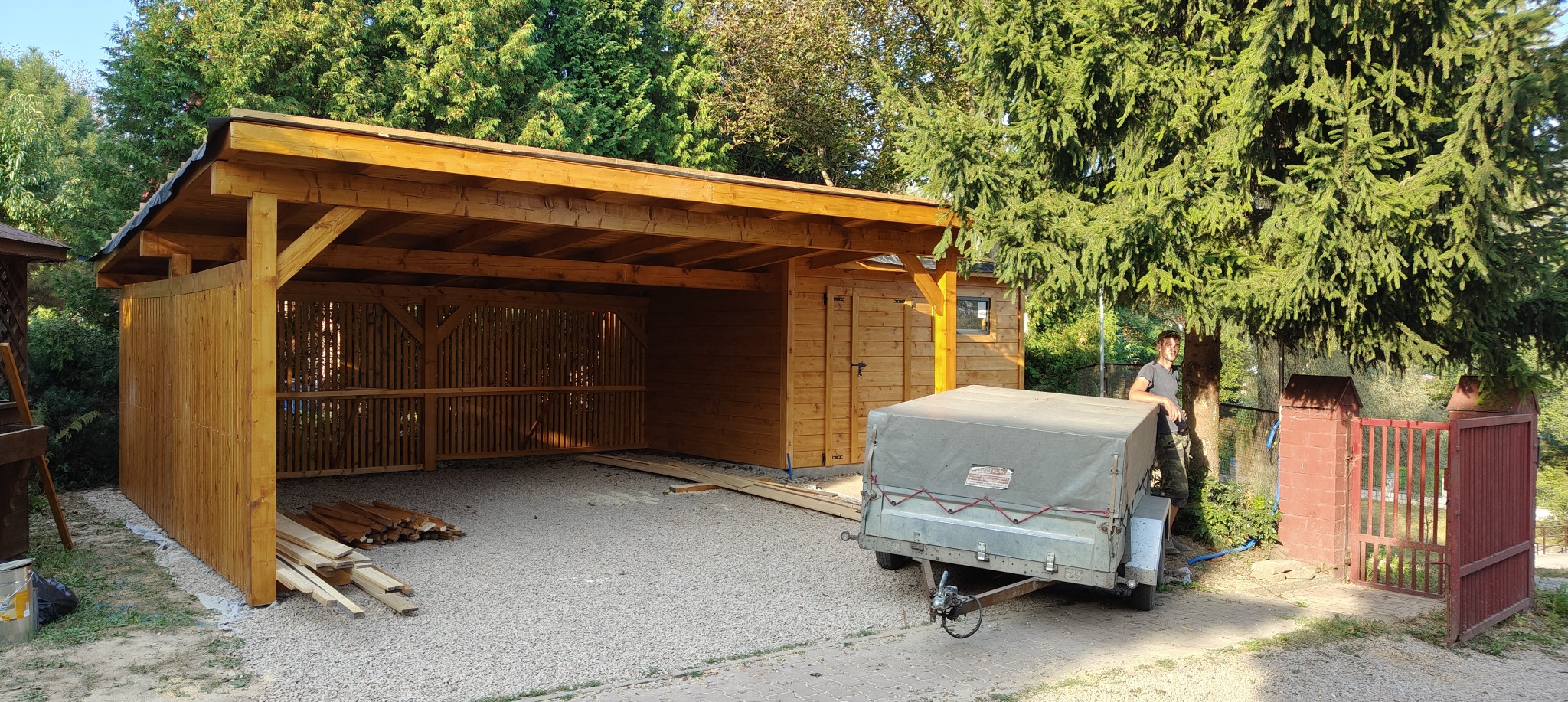 Drewniana konstrukcja z zadaszeniem, prawdopodobnie wiata garażowa lub część większej konstrukcji, z elementami dekoracyjnymi, widoczne deski i przyczepka z plandeką obok, w tle zieleń drzew.