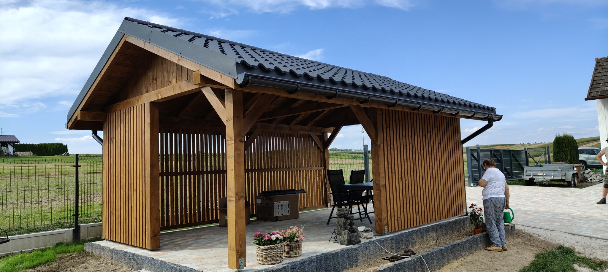 Drewniana altana z czarnym dachem, pionowe deski boczne, stół i krzesła w środku, kobieta podlewa kwiaty obok, widok na pola w tle.
