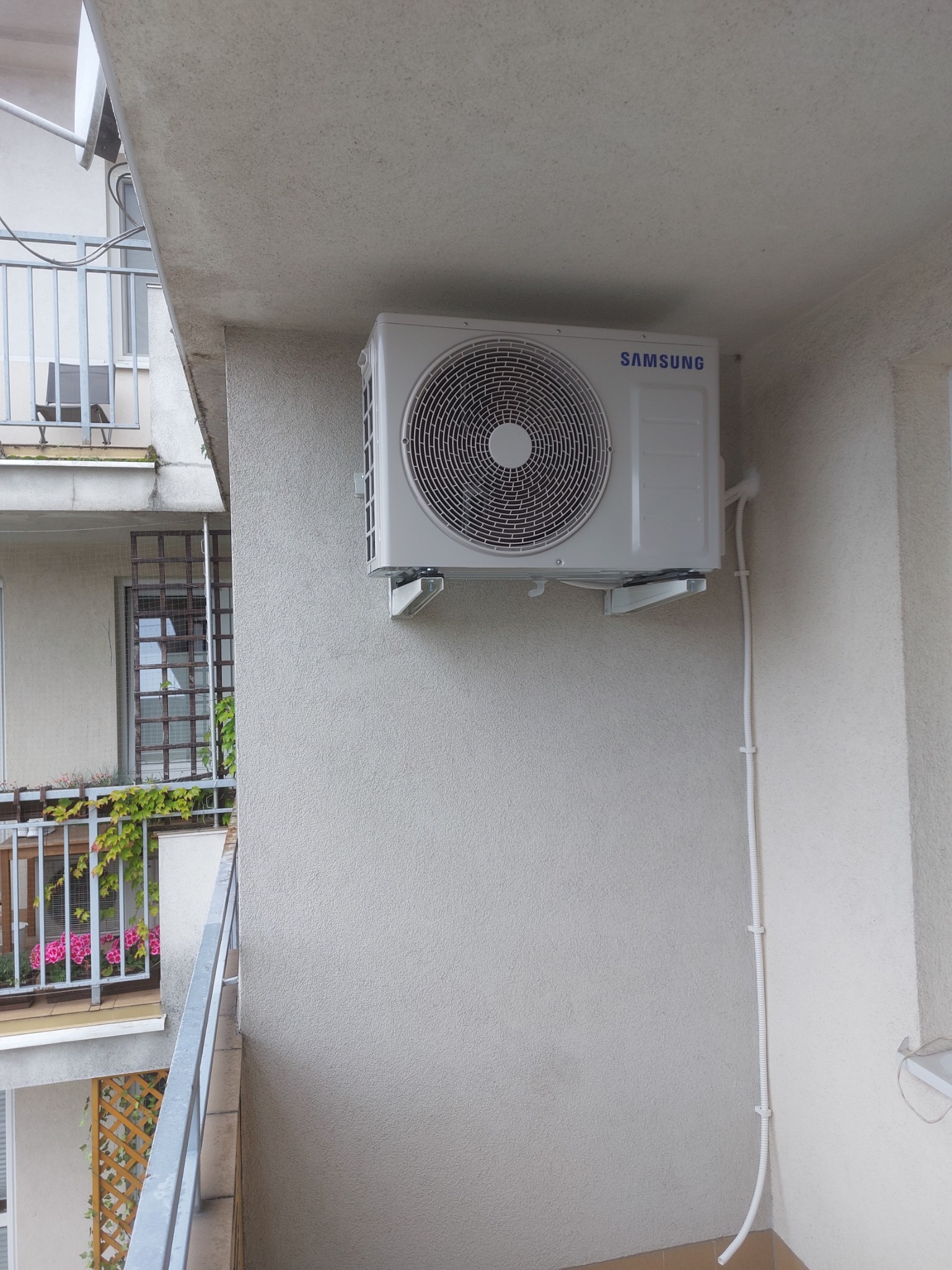 Zewnętrzna jednostka klimatyzatora Samsung zamontowana na ścianie balkonu, widoczne przewody w listwach, fragment balkonu z kwiatami w tle.