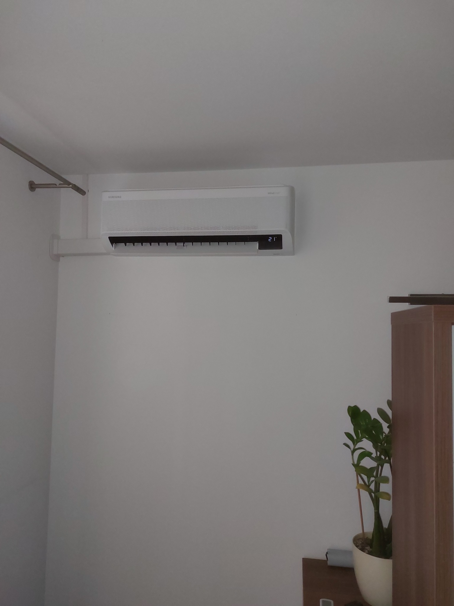 Biały klimatyzator Samsung WindFree zamontowany na ścianie w jasnym pomieszczeniu, widoczny wyświetlacz z temperaturą 21 stopni Celsjusza, obok fragment szafki i doniczka z zieloną rośliną.