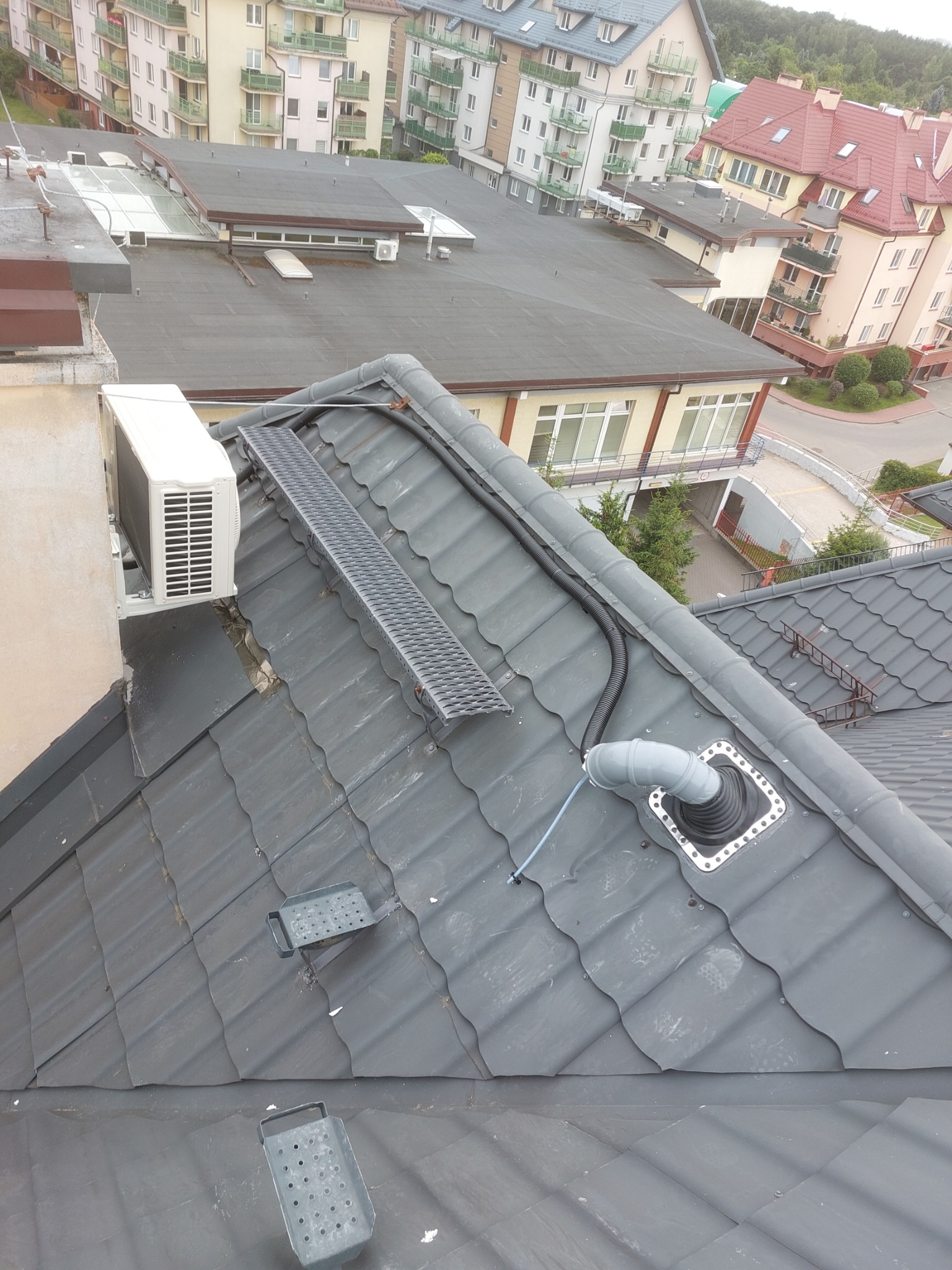 Biała jednostka zewnętrzna klimatyzacji zamontowana na dachu budynku z szarej dachówki, widoczne elementy instalacji, kratka pomostowa i komin wentylacyjny.