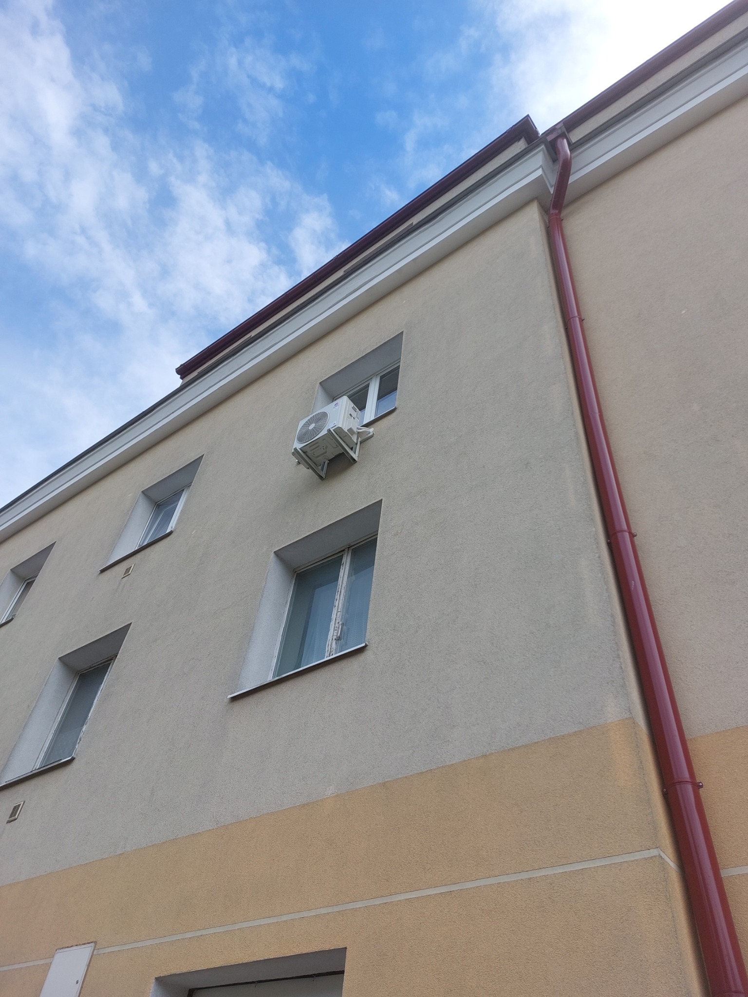 Zewnętrzna jednostka klimatyzatora zamontowana na ścianie budynku, widoczna z dołu pod kątem, z widocznym niebem w tle i rynną odprowadzającą wodę.