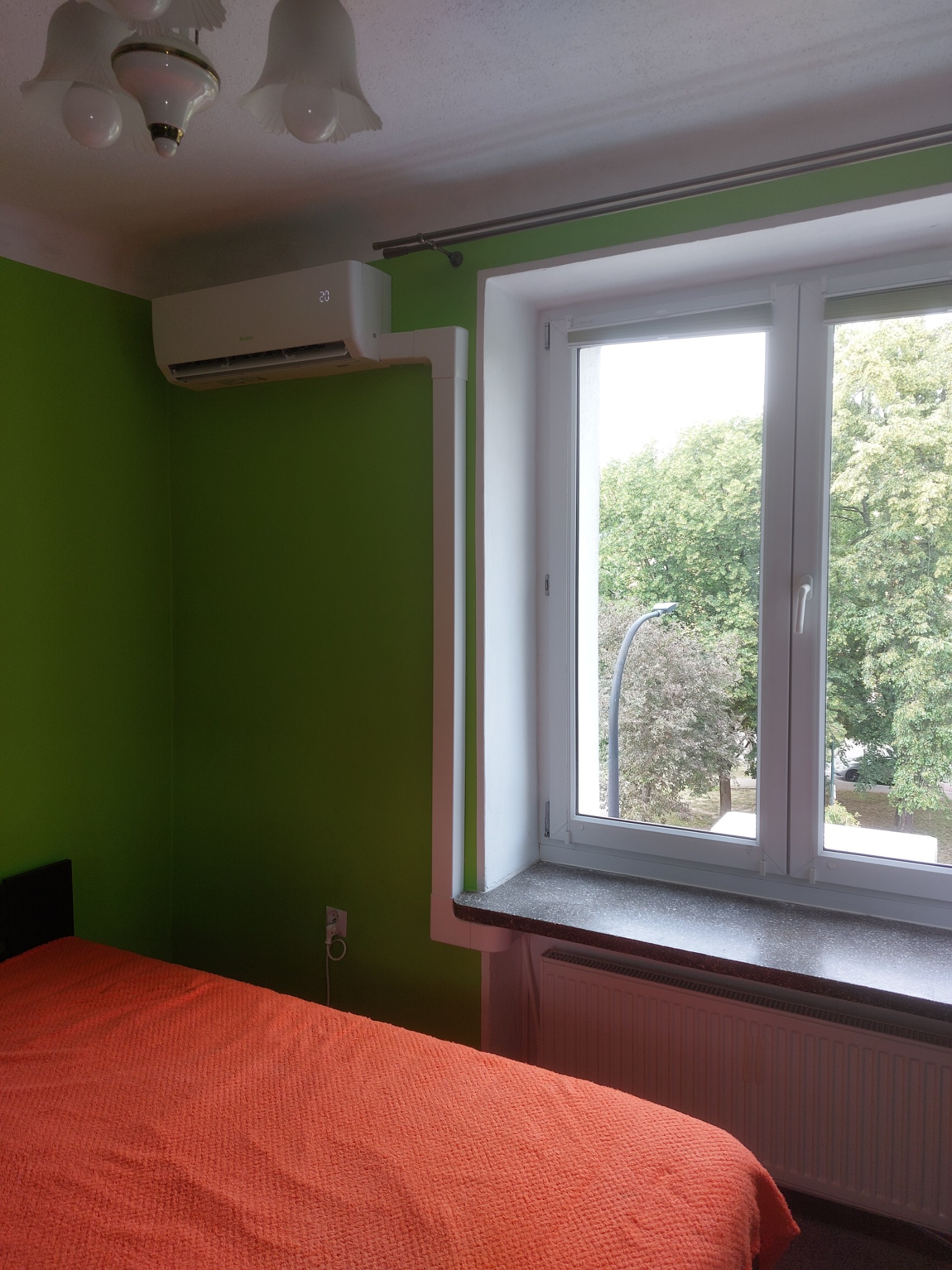 Wnętrze pokoju z zieloną ścianą, gdzie zamontowano klimatyzator ścienny nad łóżkiem, widok z okna na zieleń drzew.