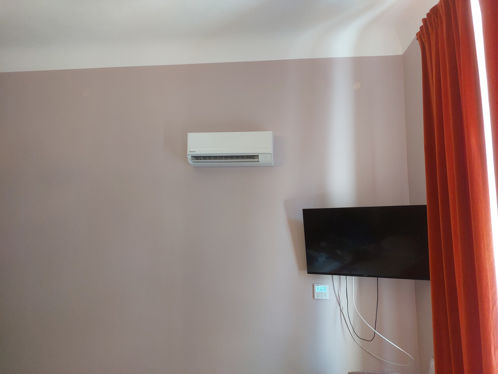 Biały klimatyzator Panasonic zamontowany na ścianie nad telewizorem z widocznym panelem sterowania, obok pomarańczowe zasłony.