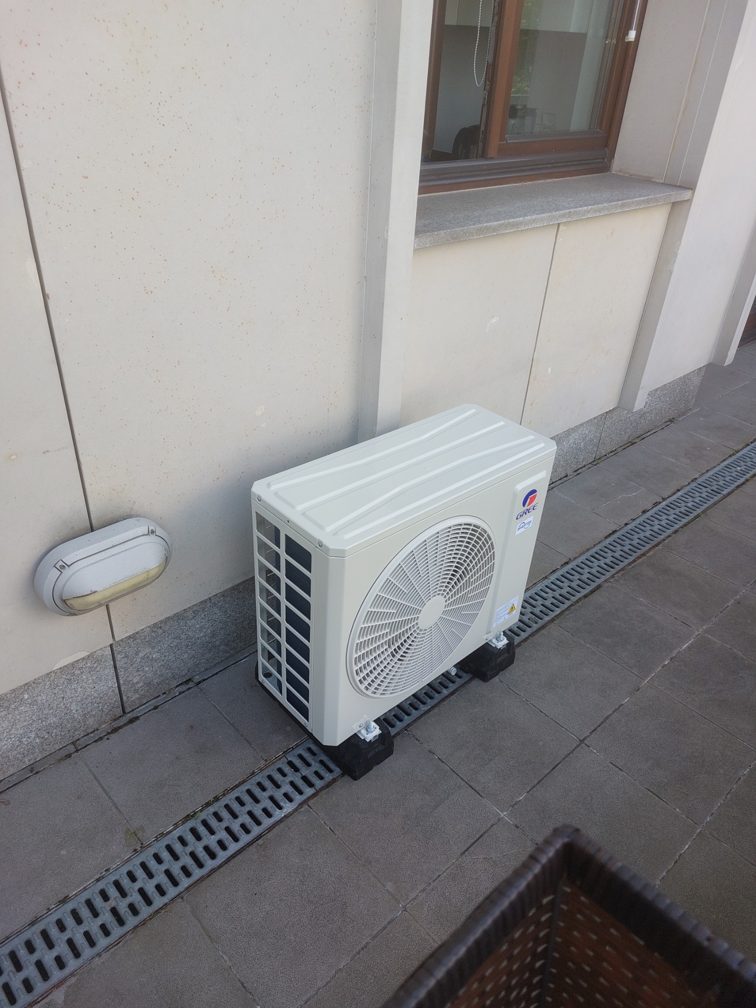 Zewnętrzna jednostka klimatyzacji marki Gree zamontowana na czarnych podkładkach antywibracyjnych przy ścianie budynku z oknem i lampą zewnętrzną, widoczna kratka odpływowa na posadzce.