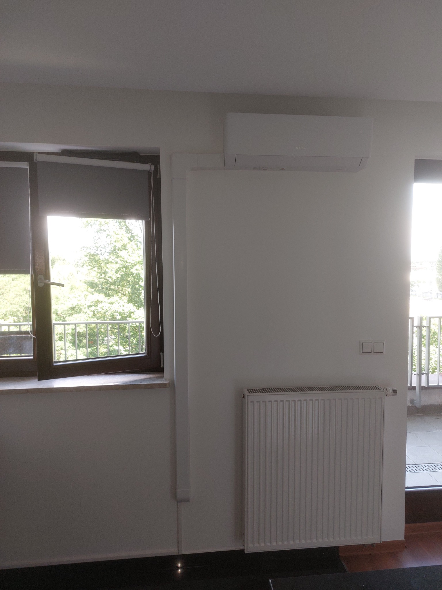 Biały klimatyzator ścienny zamontowany nad grzejnikiem w jasnym pokoju z oknem i balkonem.