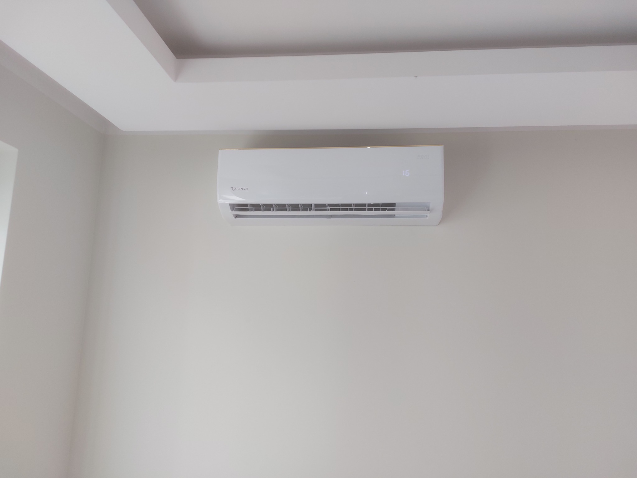 Biały klimatyzator marki Rotenso zamontowany na jasnej ścianie w pomieszczeniu z białym sufitem podwieszanym.