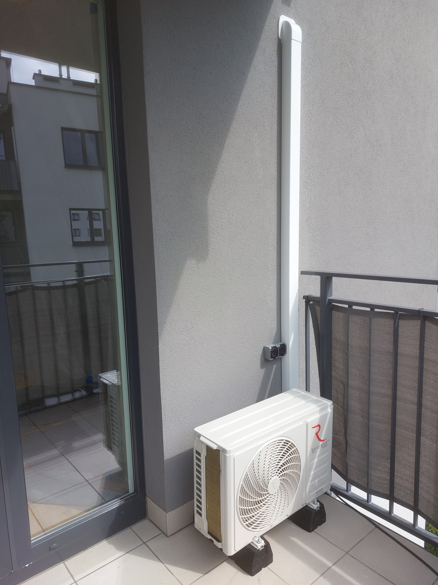 Biała jednostka zewnętrzna klimatyzacji Rotenso zamontowana na balkonie bloku mieszkalnego, widoczna rura odprowadzająca skropliny.