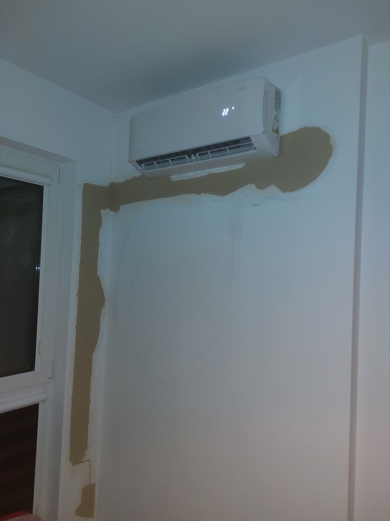 Biały klimatyzator ścienny z wyświetlaczem 22 stopnie Celsjusza, świeżo zamontowany na białej ścianie z widocznymi śladami po montażu i niedokończonymi pracami malarskimi.