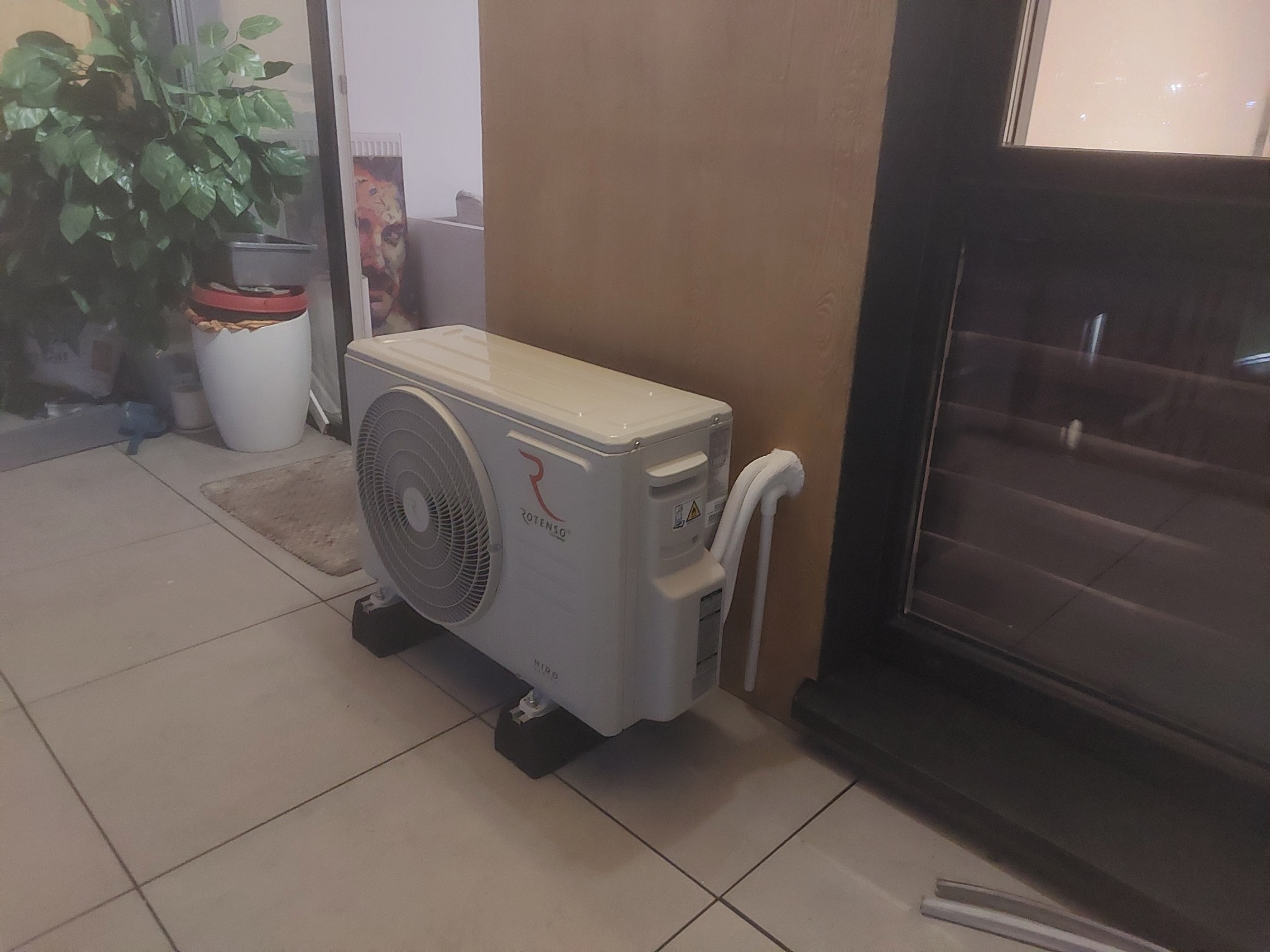 Biała, zewnętrzna jednostka klimatyzacji marki Rotenso, ustawiona na czarnych podkładkach, z widocznymi rurami odprowadzającymi, na tle beżowej ściany i ciemnej ramy okiennej, na jasnej podłodze...