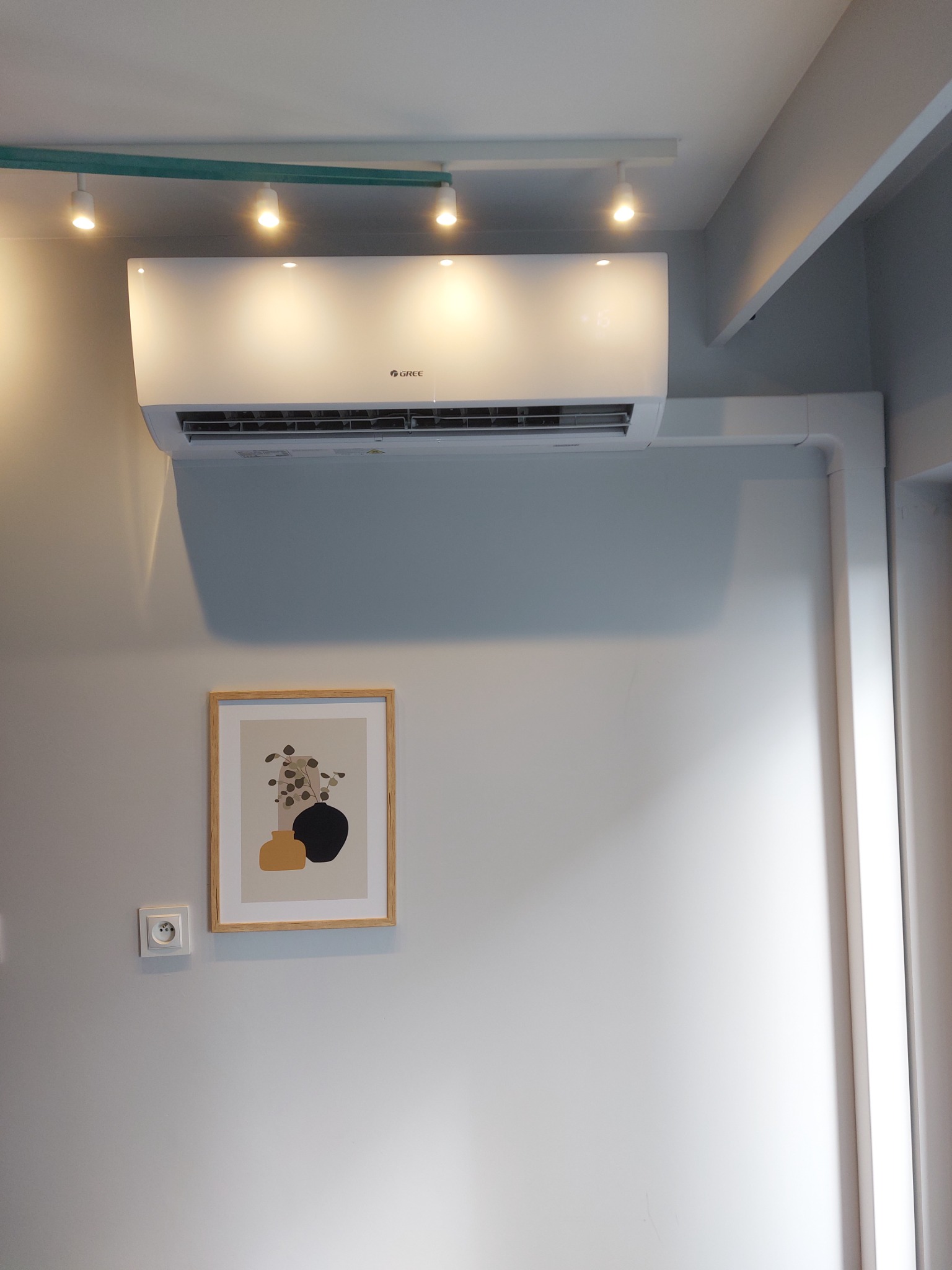 Biały klimatyzator marki Gree zamontowany na szarej ścianie w pomieszczeniu z minimalistycznym wystrojem, widoczne oświetlenie punktowe i obrazek w ramce.