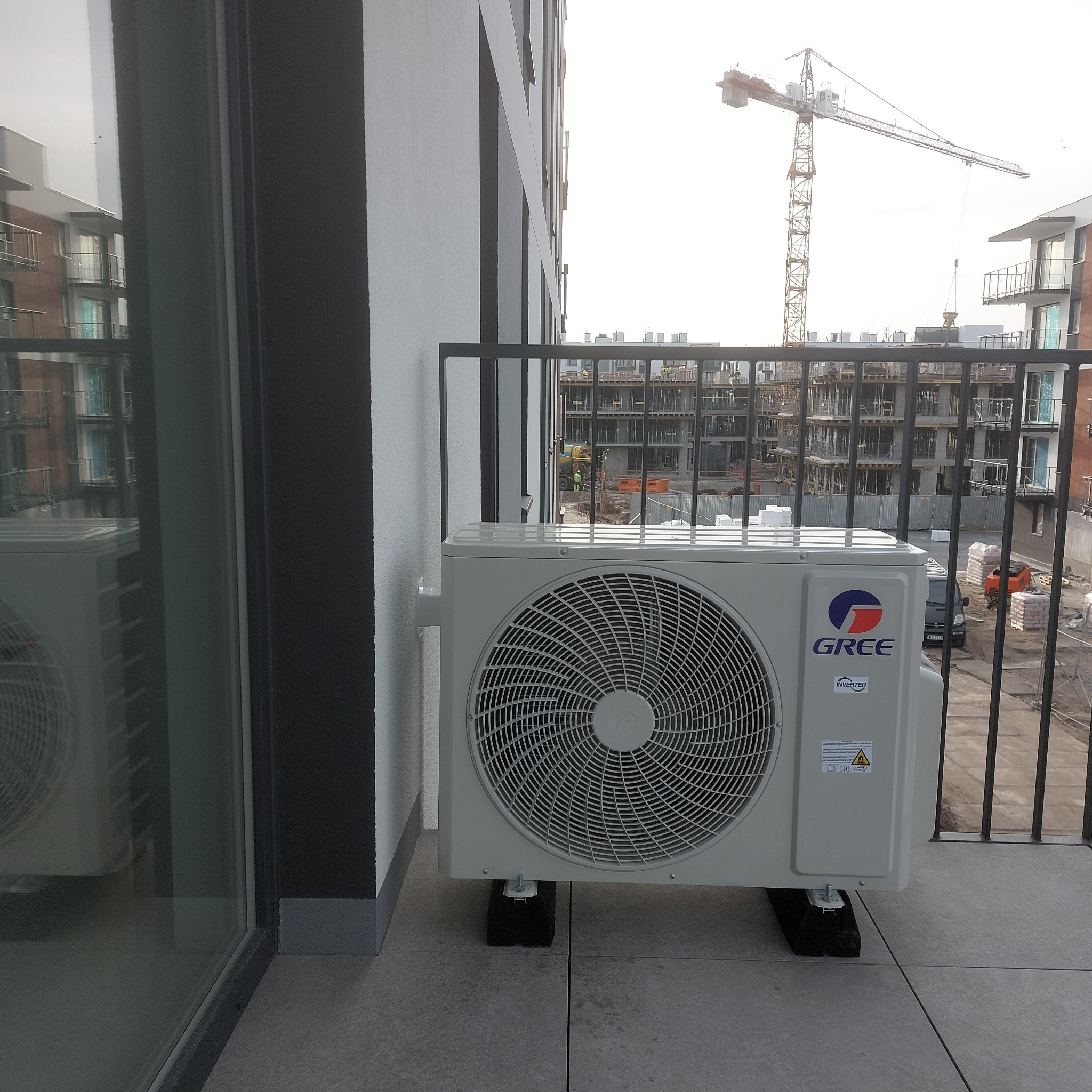 Zewnętrzna jednostka klimatyzacji marki Gree zamontowana na balkonie nowego budynku, z widokiem na plac budowy i dźwig w tle.